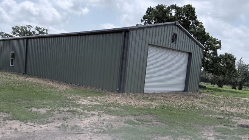 Wide view of a modern metal building with a roll-up door and gravel drive located on a rural Texas property under a cloudy sky.