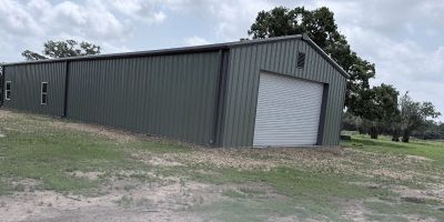 Wide view of a modern metal building with a roll-up door and gravel drive located on a rural Texas property under a cloudy sky.